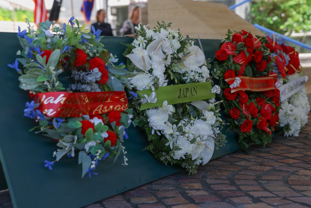 Three memorial wreaths are displayed on a green surface at what appears to be a commemorative ceremony. The center wreath bears a green ribbon labeled 'JAPAN', while the left wreath has a red ribbon with partially visible text referencing what appears to be a Hawaiian veterans or merchant association. The right wreath features red roses with a partially visible ribbon, and two individuals are visible in the blurred background.