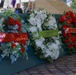 Three memorial wreaths are displayed on a green surface at what appears to be a commemorative ceremony. The center wreath bears a green ribbon labeled 'JAPAN', while the left wreath has a red ribbon with partially visible text referencing what appears to be a Hawaiian veterans or merchant association. The right wreath features red roses with a partially visible ribbon, and two individuals are visible in the blurred background.