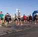 A large group of runners is crossing or departing from a finish line arch at what appears to be a military base road race. The blue inflatable 'FINISH' arch is visible on the right side, and participants are dressed in casual athletic wear. The event takes place on a paved surface with military hangar buildings and a light tower visible in the background.