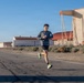 A runner is captured mid-stride on what appears to be an airfield or runway, approaching a finish line marked by a blue inflatable arch. The background features large industrial or aerospace hangars, including a distinctive structure with a red curved roof and metal scaffolding framework. The setting suggests this is an organized race event held on a military or government airfield.