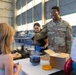 Two U.S. Air Force personnel are conducting a science demonstration for children inside what appears to be a military hangar. One airman, whose name tag reads 'BELL,' is smiling and engaging with young attendees while a colleague handles what appears to be a dry ice or chemical experiment. The event appears to be a STEM outreach or community engagement activity, evidenced by the rocket balloon decoration and science materials on the table.