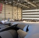A large military or aviation hangar hosts a public open house or air show event, with a massive multi-engine aircraft (likely a C-5 Galaxy or C-17 Globemaster) visible in the foreground. A large American flag is prominently displayed on the hangar wall. Crowds of visitors browse vendor tents and exhibits on the hangar floor below.