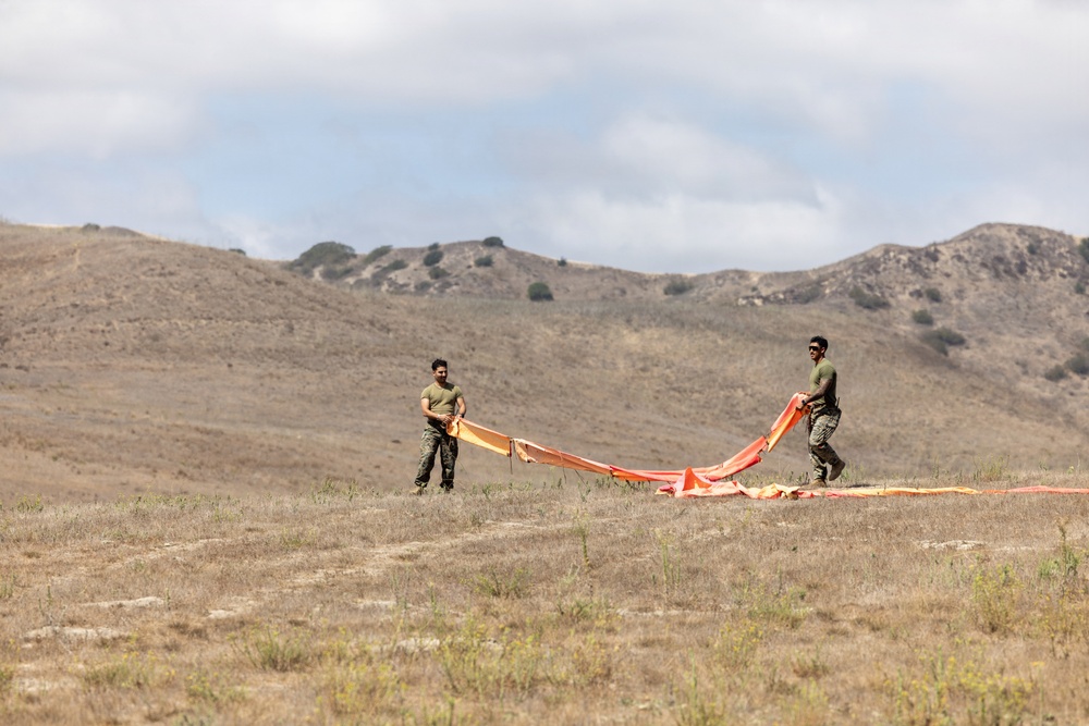 Two individuals in military-style olive green uniforms are handling a large orange fabric panel or signal marker on an open, dry grassland field. They appear to be spreading or folding the material across the ground. The setting features arid rolling hills in the background under a partly cloudy sky.