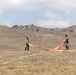 Two individuals in military-style olive green uniforms are handling a large orange fabric panel or signal marker on an open, dry grassland field. They appear to be spreading or folding the material across the ground. The setting features arid rolling hills in the background under a partly cloudy sky.