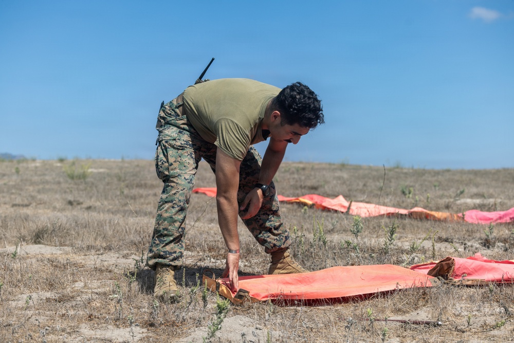 A military service member in MARPAT camouflage uniform and olive drab t-shirt is bent over arranging or placing an orange panel marker on dry, sparse terrain. Multiple additional orange panels are visible in the background, suggesting a landing zone or drop zone marking operation. The setting appears to be an open field or training area under clear blue skies.