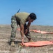 A military service member in MARPAT camouflage uniform and olive drab t-shirt is bent over arranging or placing an orange panel marker on dry, sparse terrain. Multiple additional orange panels are visible in the background, suggesting a landing zone or drop zone marking operation. The setting appears to be an open field or training area under clear blue skies.