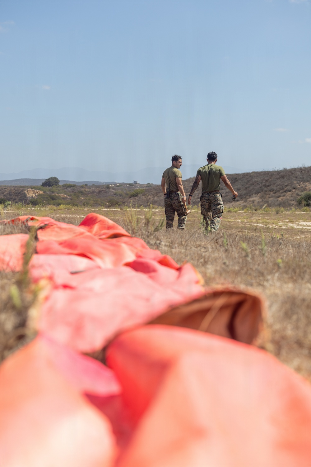 Two military personnel in camouflage pants and olive green t-shirts stand in an open field, viewed from behind. In the foreground, a large orange parachute or canopy lies deflated on the dry grass. The setting appears to be an arid training area with scrubland and hills visible in the background.