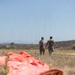 Two military personnel in camouflage pants and olive green t-shirts stand in an open field, viewed from behind. In the foreground, a large orange parachute or canopy lies deflated on the dry grass. The setting appears to be an arid training area with scrubland and hills visible in the background.