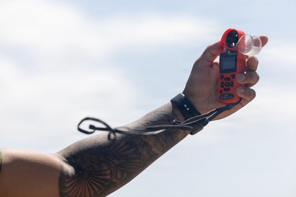 A tattooed hand and forearm are raised against an overcast sky, holding a red Kestrel weather meter/anemometer. The device features a small impeller fan at the top, a digital display screen, and multiple buttons. A black wrist strap or lanyard is attached to the device and wrapped around the wrist.