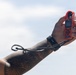 A tattooed hand and forearm are raised against an overcast sky, holding a red Kestrel weather meter/anemometer. The device features a small impeller fan at the top, a digital display screen, and multiple buttons. A black wrist strap or lanyard is attached to the device and wrapped around the wrist.