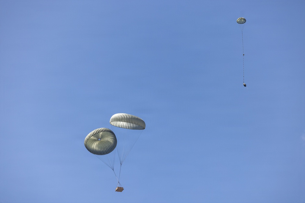 The image shows multiple military-style cargo parachutes descending against a clear blue sky. At least three parachutes are visible at varying altitudes, each carrying suspended cargo loads beneath them. The parachutes appear to be olive/sage green in color, consistent with military airdrop operations.