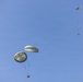 The image shows multiple military-style cargo parachutes descending against a clear blue sky. At least three parachutes are visible at varying altitudes, each carrying suspended cargo loads beneath them. The parachutes appear to be olive/sage green in color, consistent with military airdrop operations.