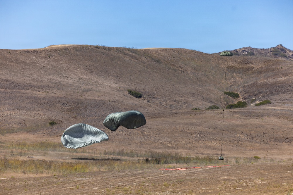 The image shows a military airdrop operation taking place over an arid, hilly landscape. Multiple olive-drab/gray cargo parachutes are visible at different stages of descent, with at least two near ground level and one still airborne at higher altitude. A drop zone marker (red panel) is visible on the ground, indicating a designated landing area.