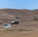 The image shows a military airdrop operation taking place over an arid, hilly landscape. Multiple olive-drab/gray cargo parachutes are visible at different stages of descent, with at least two near ground level and one still airborne at higher altitude. A drop zone marker (red panel) is visible on the ground, indicating a designated landing area.