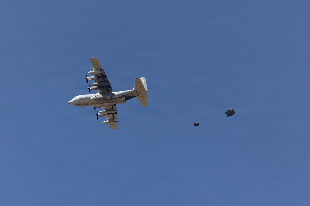 A military transport aircraft, consistent with a C-130 Hercules, is photographed from below against a clear blue sky. The aircraft is performing an airdrop operation, with a cargo bundle visibly deployed and connected to an extraction parachute trailing behind the aircraft. The image captures the moment shortly after the cargo has exited the rear of the aircraft.