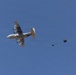 A military transport aircraft, consistent with a C-130 Hercules, is photographed from below against a clear blue sky. The aircraft is performing an airdrop operation, with a cargo bundle visibly deployed and connected to an extraction parachute trailing behind the aircraft. The image captures the moment shortly after the cargo has exited the rear of the aircraft.