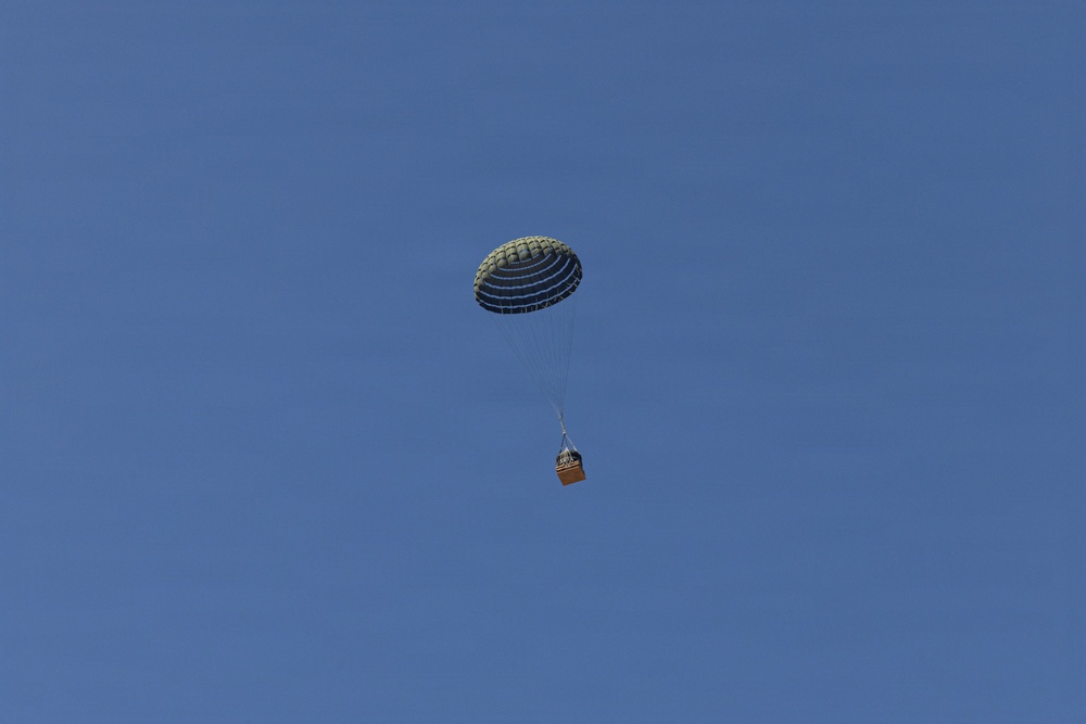 A military-style cargo parachute is descending through a clear blue sky, carrying a small brown/orange crate or box suspended beneath it by suspension lines. The parachute appears to be an olive-drab or dark green round canopy, consistent with military airdrop equipment. The image captures a supply or equipment drop in progress with no other objects or people visible.