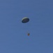 A military-style cargo parachute is descending through a clear blue sky, carrying a small brown/orange crate or box suspended beneath it by suspension lines. The parachute appears to be an olive-drab or dark green round canopy, consistent with military airdrop equipment. The image captures a supply or equipment drop in progress with no other objects or people visible.