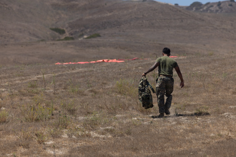 A military service member wearing a green t-shirt and camouflage pants walks across a dry, arid field carrying military gear. In the background, orange/red panels or markers are laid out on the ground, likely used as landing zone or drop zone markers. The terrain appears to be a training area with rolling hills and sparse dry vegetation.