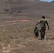 A military service member wearing a green t-shirt and camouflage pants walks across a dry, arid field carrying military gear. In the background, orange/red panels or markers are laid out on the ground, likely used as landing zone or drop zone markers. The terrain appears to be a training area with rolling hills and sparse dry vegetation.