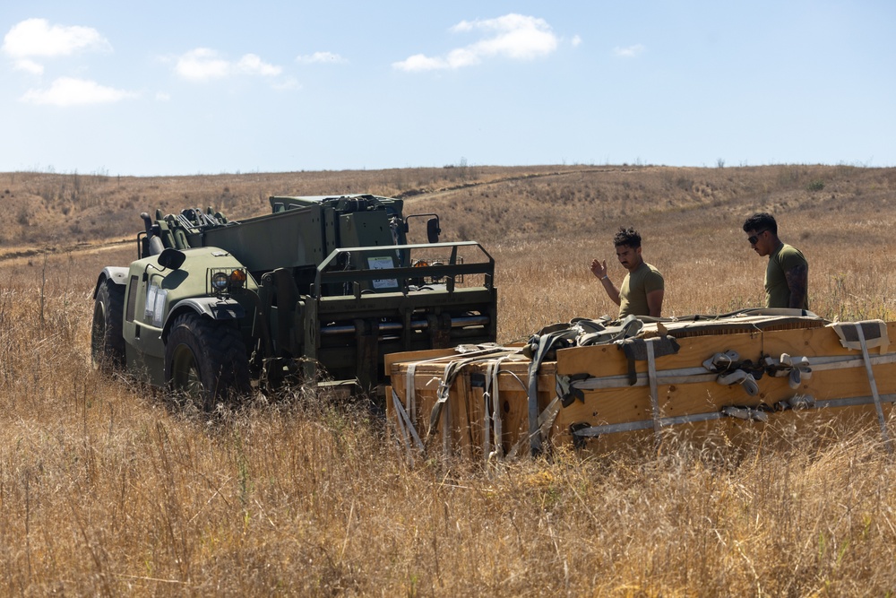 Two military personnel in olive drab t-shirts are working in a dry, grassy field alongside a military rough-terrain forklift (likely a Gradall or similar telehandler). They appear to be managing wooden crates or pallets with strapping, possibly unloading or positioning cargo. The setting appears to be a training area or field operation during daytime under clear skies.
