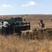 Two military personnel in olive drab t-shirts are working in a dry, grassy field alongside a military rough-terrain forklift (likely a Gradall or similar telehandler). They appear to be managing wooden crates or pallets with strapping, possibly unloading or positioning cargo. The setting appears to be a training area or field operation during daytime under clear skies.