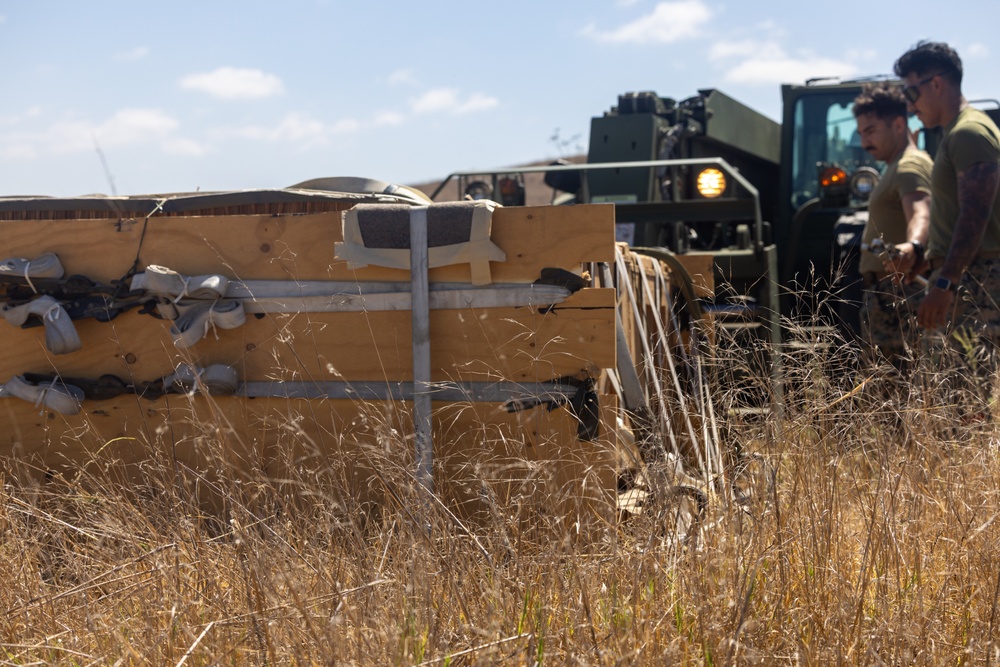 Military personnel are working in a dry, grassy field alongside a military forklift or rough-terrain vehicle. A large wooden crate or packing container is visible in the foreground, partially opened or damaged, with straps and padding materials visible. The scene appears to depict a logistics or equipment unpacking operation in an outdoor field environment.