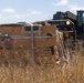 Military personnel are working in a dry, grassy field alongside a military forklift or rough-terrain vehicle. A large wooden crate or packing container is visible in the foreground, partially opened or damaged, with straps and padding materials visible. The scene appears to depict a logistics or equipment unpacking operation in an outdoor field environment.