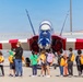 A group of school-aged children on a field trip are gathered on an airfield tarmac, viewing a NASA-marked jet aircraft up close. An adult chaperone or educator stands with the group, appearing to point out features of the aircraft. The jet is painted in a distinctive red, white, and blue color scheme with wing markings.