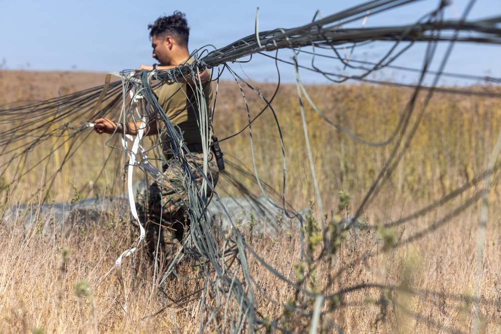 A person wearing military camouflage uniform is working in an open field with dry grassland vegetation. They are handling a large bundle of tangled wires or cables, appearing to either deploy or recover the material. The setting appears to be a rural or training area under clear sky conditions.