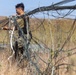 A person wearing military camouflage uniform is working in an open field with dry grassland vegetation. They are handling a large bundle of tangled wires or cables, appearing to either deploy or recover the material. The setting appears to be a rural or training area under clear sky conditions.