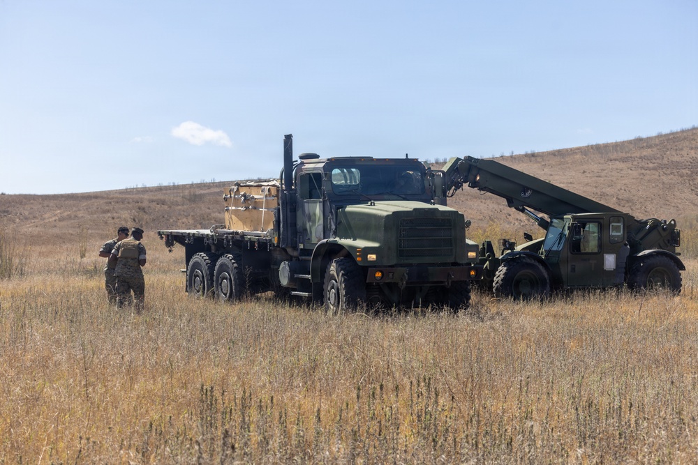 A military field operation is underway in an open, dry grassland area. Two large military tactical trucks are positioned in the field, with at least one soldier in camouflage uniform standing nearby. The scene appears to depict a logistics or equipment handling exercise.