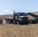 A military field operation is underway in an open, dry grassland area. Two large military tactical trucks are positioned in the field, with at least one soldier in camouflage uniform standing nearby. The scene appears to depict a logistics or equipment handling exercise.