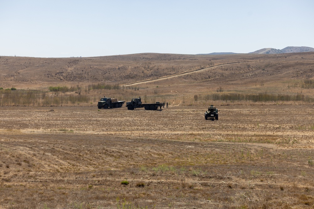 A wide open arid landscape showing military vehicles and personnel operating in a flat, dry field. Multiple trucks and at least one armored or wheeled military vehicle are visible in the mid-ground, with rolling brown hills in the background. The scene appears to depict a military field operation or exercise in a semi-arid environment.