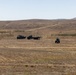 A wide open arid landscape showing military vehicles and personnel operating in a flat, dry field. Multiple trucks and at least one armored or wheeled military vehicle are visible in the mid-ground, with rolling brown hills in the background. The scene appears to depict a military field operation or exercise in a semi-arid environment.