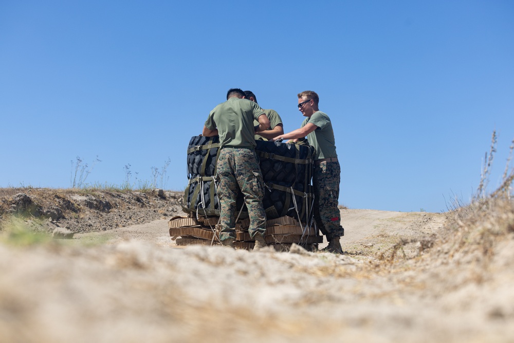 Three military personnel in olive green t-shirts and camouflage trousers are working together to handle what appears to be airdrop or parachute equipment on arid, sandy terrain. The individuals are wearing uniforms consistent with U.S. Marine Corps digital camouflage (MARPAT) pattern trousers. The scene takes place outdoors under a clear blue sky in a dry, open landscape.