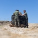 Three military personnel in olive green t-shirts and camouflage trousers are working together to handle what appears to be airdrop or parachute equipment on arid, sandy terrain. The individuals are wearing uniforms consistent with U.S. Marine Corps digital camouflage (MARPAT) pattern trousers. The scene takes place outdoors under a clear blue sky in a dry, open landscape.