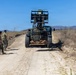 Military personnel in olive drab uniforms are operating a rough-terrain forklift (likely a military RTFL/telehandler) on a dirt road in an arid, hilly training environment. The forklift is carrying a loaded pallet secured with straps, and three service members are positioned around the vehicle, appearing to guide or supervise the operation. The setting is consistent with a U.S. Marine Corps training base, possibly Camp Pendleton, California.