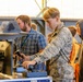 A group of young students, one wearing a military camouflage uniform, are participating in what appears to be a STEM educational event or air show demonstration. A bearded adult volunteer in a plaid shirt with a lanyard is supervising as the uniformed youth operates a flight simulator joystick in front of an LG monitor. A scale model aircraft (resembling an F-22 or similar stealth fighter) is mounted on the display station.