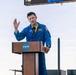 A NASA astronaut in a blue flight suit is speaking at a podium bearing the NASA logo at an outdoor event. An American flag with gold fringe is visible to the right of the speaker. The setting appears to be a formal NASA public address or pre-launch event.