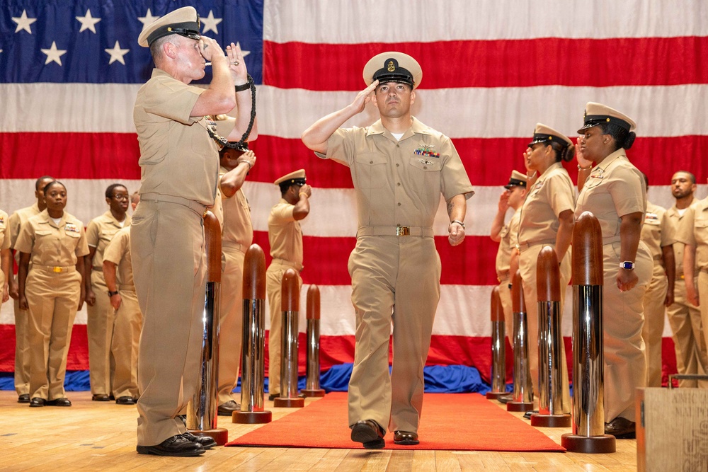 A formal U.S. Navy military ceremony is taking place in front of a large American flag backdrop. A central figure in khaki service uniform walks along a red carpet between chrome stanchions while exchanging salutes with a senior officer. Rows of Navy personnel in matching uniforms stand at attention on both sides.