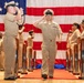A formal U.S. Navy military ceremony is taking place in front of a large American flag backdrop. A central figure in khaki service uniform walks along a red carpet between chrome stanchions while exchanging salutes with a senior officer. Rows of Navy personnel in matching uniforms stand at attention on both sides.