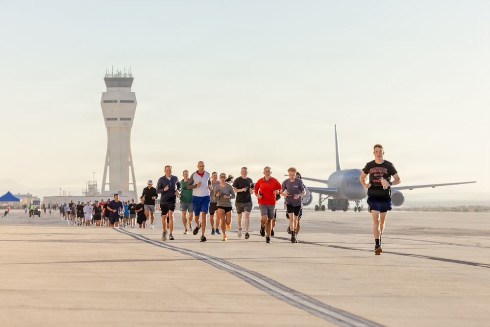 A large group of people are running on an airport tarmac or runway during what appears to be an organized run or race event. An air traffic control tower is visible in the background on the left, and a large commercial or military aircraft is parked on the right side. The lighting suggests early morning conditions, with a warm, hazy sky.