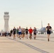 A large group of people are running on an airport tarmac or runway during what appears to be an organized run or race event. An air traffic control tower is visible in the background on the left, and a large commercial or military aircraft is parked on the right side. The lighting suggests early morning conditions, with a warm, hazy sky.