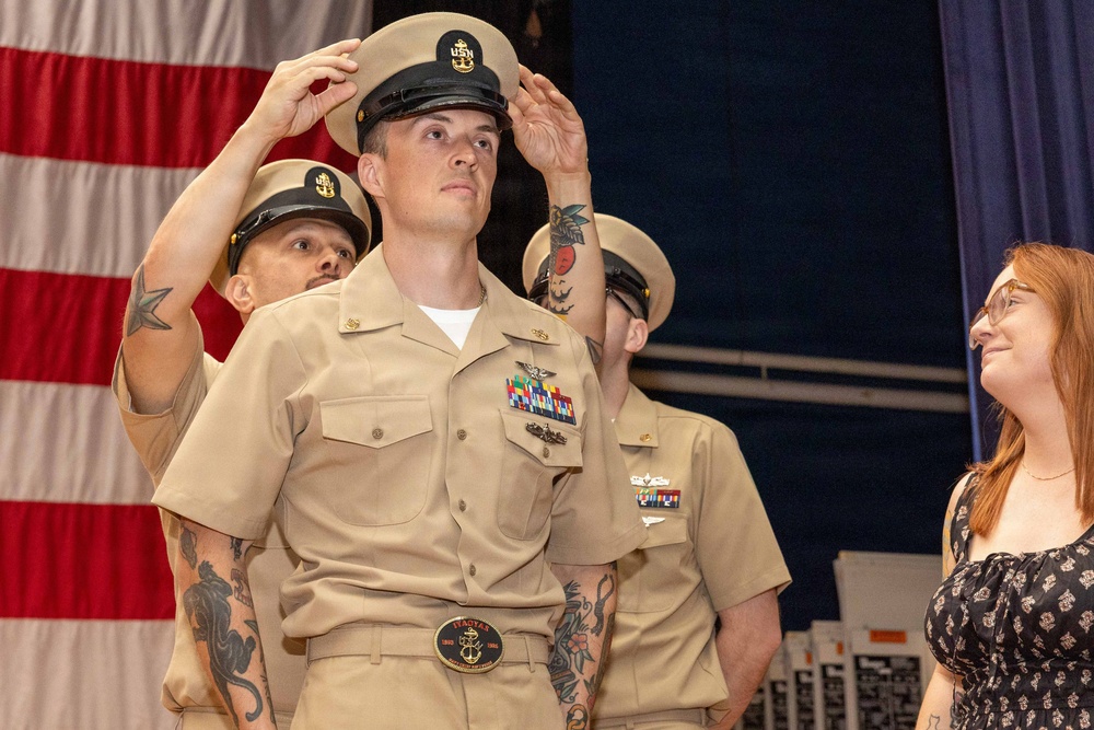 A U.S. Navy Chief Petty Officer pinning/hat ceremony is taking place, with a sailor having his chief's cover (hat) placed on his head by fellow service members. The central figure is in khaki uniform adorned with multiple ribbons, badges, and a distinctive belt buckle. An American flag is visible in the background, and a civilian woman with red hair observes the ceremony.