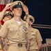 A U.S. Navy Chief Petty Officer pinning/hat ceremony is taking place, with a sailor having his chief's cover (hat) placed on his head by fellow service members. The central figure is in khaki uniform adorned with multiple ribbons, badges, and a distinctive belt buckle. An American flag is visible in the background, and a civilian woman with red hair observes the ceremony.