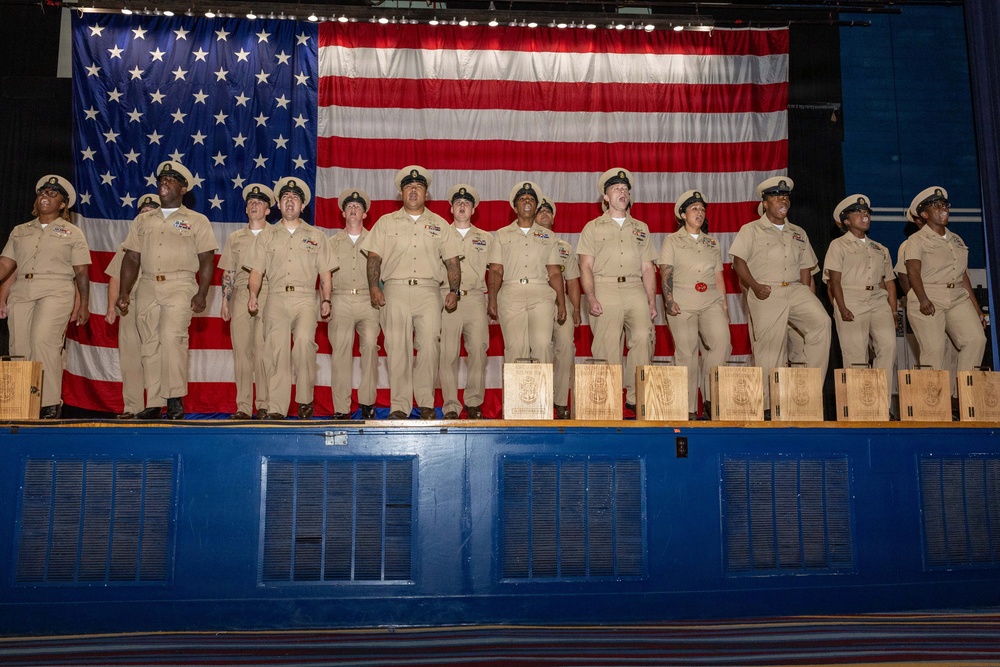A group of approximately 13 U.S. Navy personnel in khaki service uniforms stand in a line on a stage in front of a large American flag. Wooden plaques or boxes are arranged at their feet on the stage. This appears to be a formal military ceremony, likely a chief petty officer pinning or promotion ceremony.