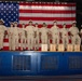 A group of approximately 13 U.S. Navy personnel in khaki service uniforms stand in a line on a stage in front of a large American flag. Wooden plaques or boxes are arranged at their feet on the stage. This appears to be a formal military ceremony, likely a chief petty officer pinning or promotion ceremony.