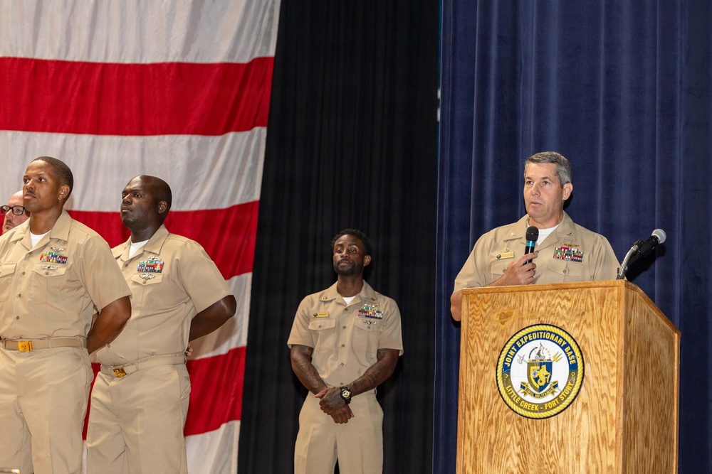 A formal military ceremony is taking place at Joint Expeditionary Base Little Creek-Fort Story, as indicated by the podium seal. A senior Navy officer in khaki service uniform is speaking at the podium while three other Navy personnel stand at attention in the background. A large American flag is displayed prominently on the left side of the stage.