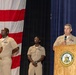 A formal military ceremony is taking place at Joint Expeditionary Base Little Creek-Fort Story, as indicated by the podium seal. A senior Navy officer in khaki service uniform is speaking at the podium while three other Navy personnel stand at attention in the background. A large American flag is displayed prominently on the left side of the stage.